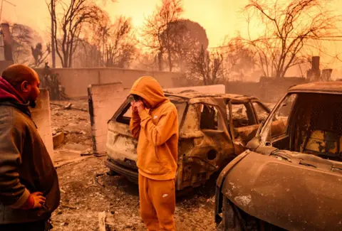 Getty Images A young man cries as he surveys the wreckage of a cars burnt out in the Los Angeles fires. Another man looks on.